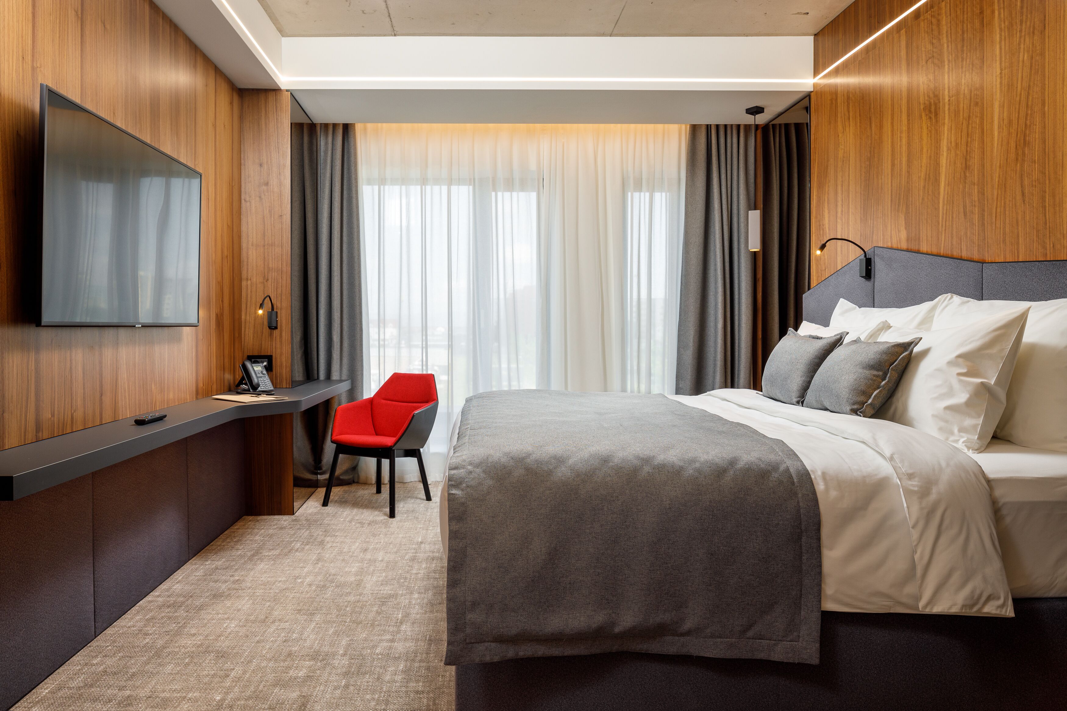 A modern hotel room with a large bed, a desk with a chair, a flat-screen TV, and a large window with curtains.