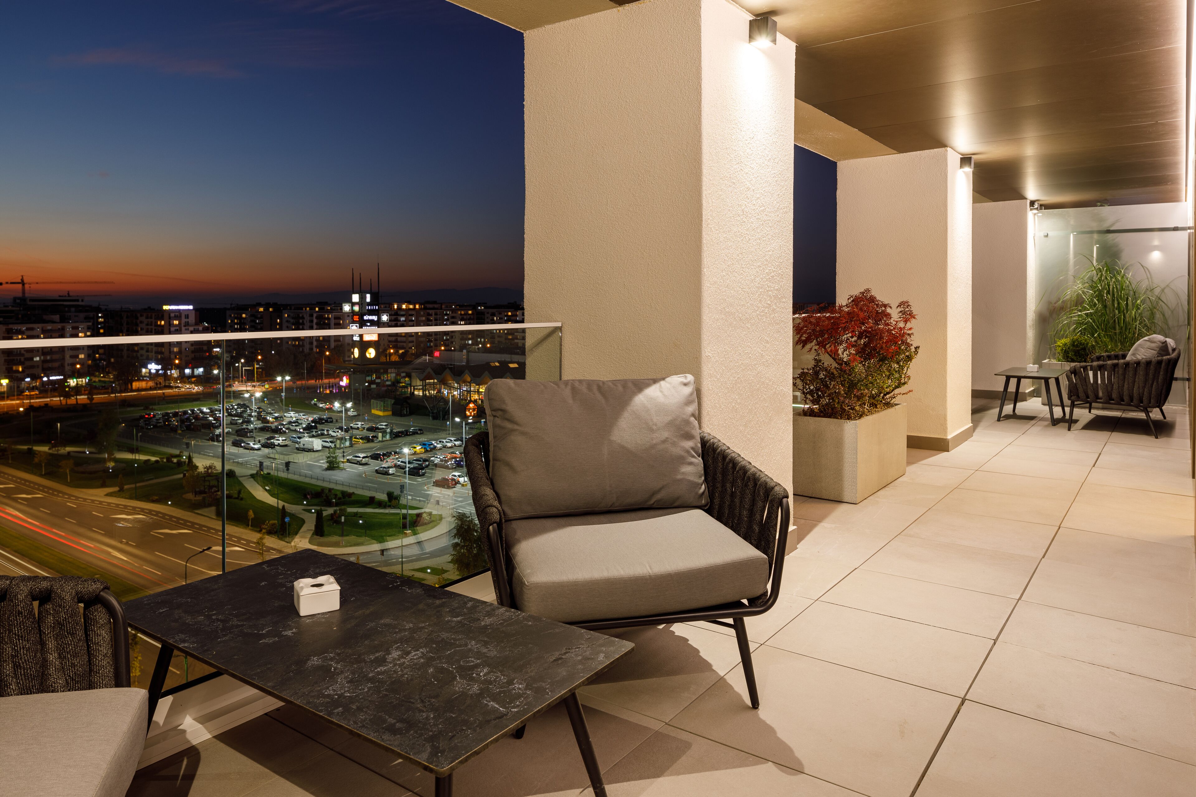A balcony with a view of a cityscape at dusk, a cushioned chair, a small table, and a potted plant.