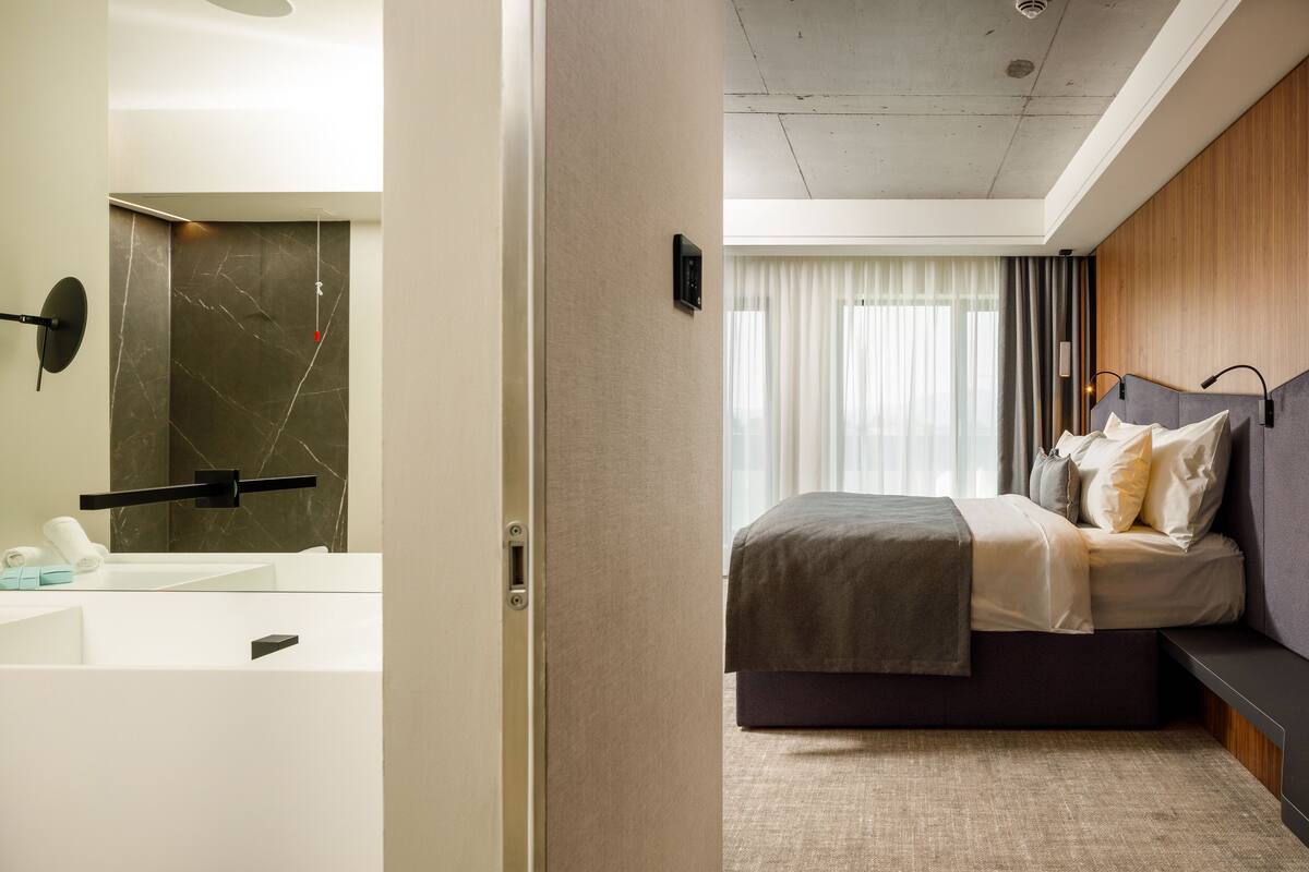 A modern hotel room with a large bed, a built-in bench, a bathtub, and a bathroom with a marble wall.