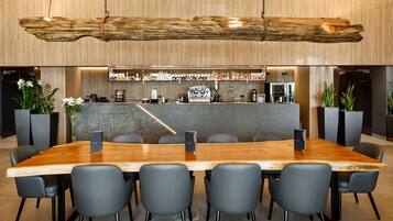 Una sala de conferencias moderna con una larga mesa de madera, sillas negras, una zona de bar con varias botellas y plantas en macetas.