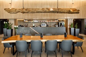 Una sala de conferencias moderna con una larga mesa de madera, sillas negras, una zona de bar con varias botellas y plantas en macetas.