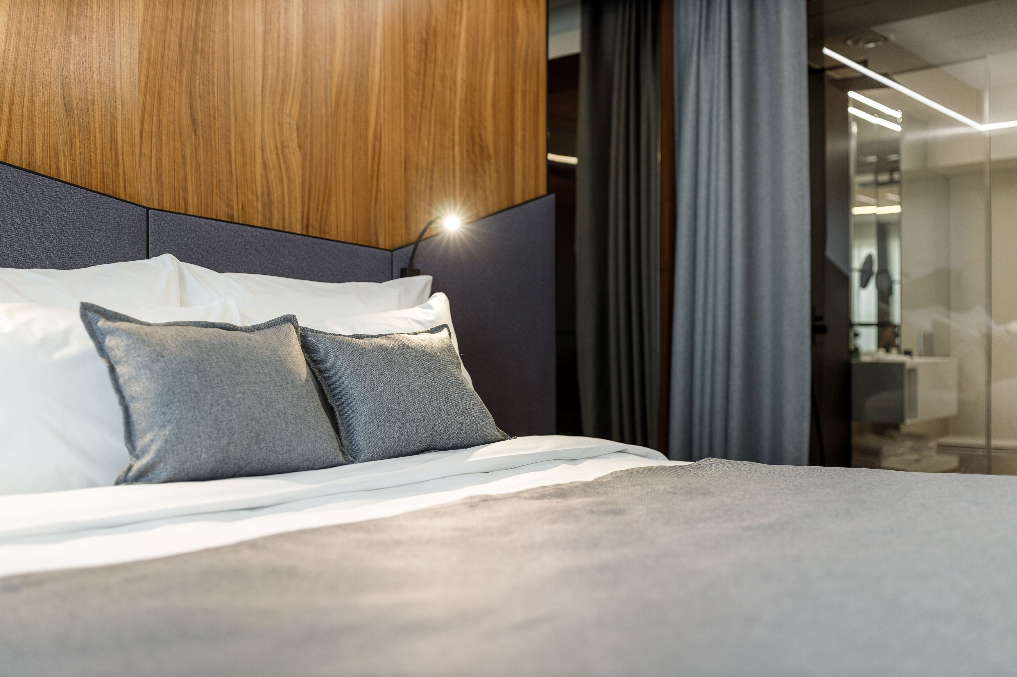 Une chambre à coucher moderne avec une tête de lit en bois, un lit recouvert de draps blancs et agrémenté d’oreillers gris.