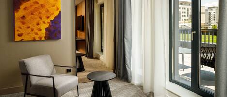 A modern hotel room with a large window, a comfortable chair, a small round table, and a colorful abstract painting on the wall.
