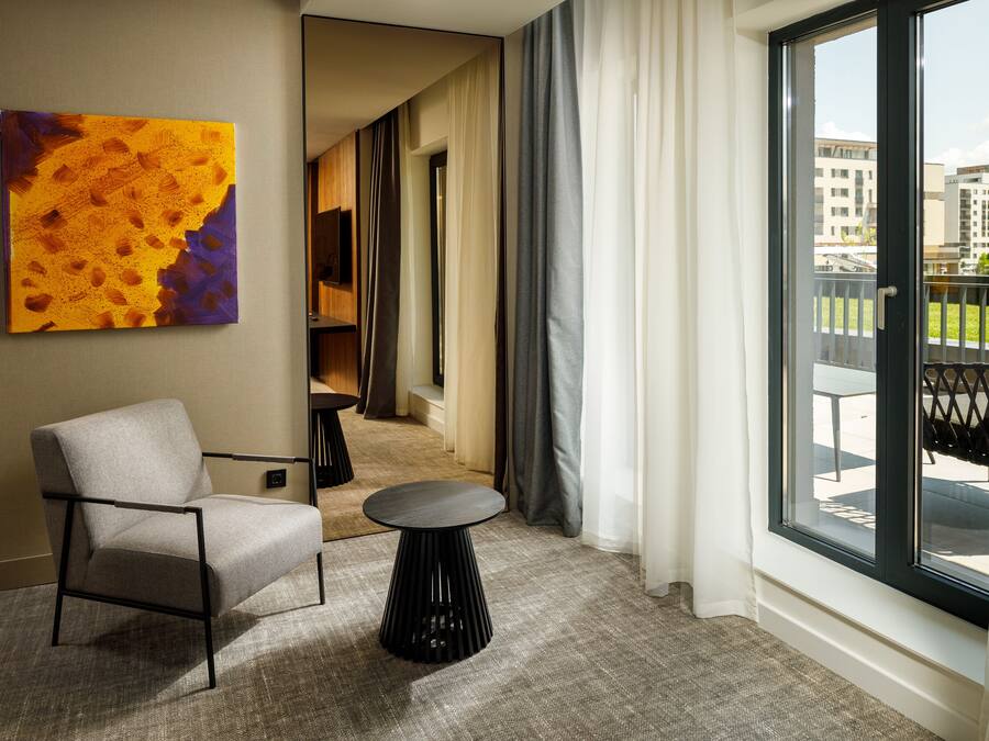 A modern hotel room with a large window, a comfortable chair, a small round table, and a colorful abstract painting on the wall.