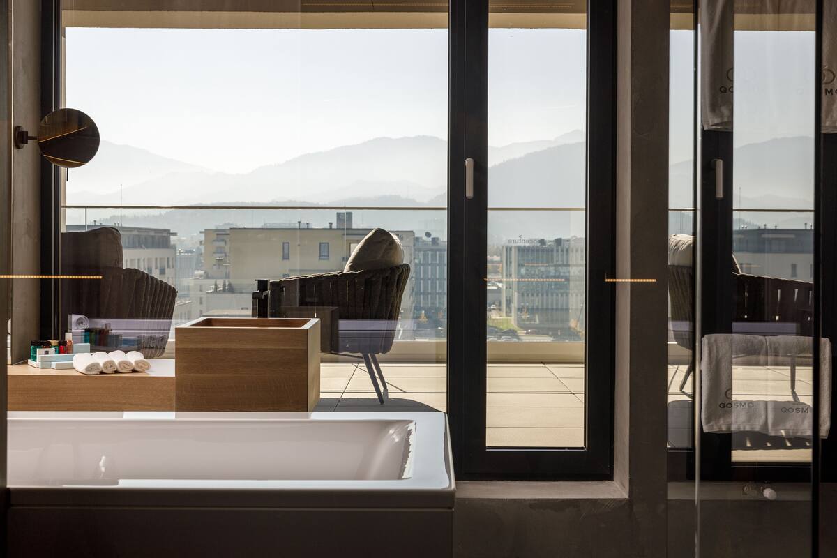 A modern hotel room with a large window, a wooden desk, a chair, and a bed.