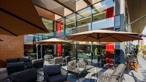 An outdoor seating area with modern furniture and large umbrellas, overlooking a glass building with red curtains. - Qosmo Brasov Hotel (Brasov)