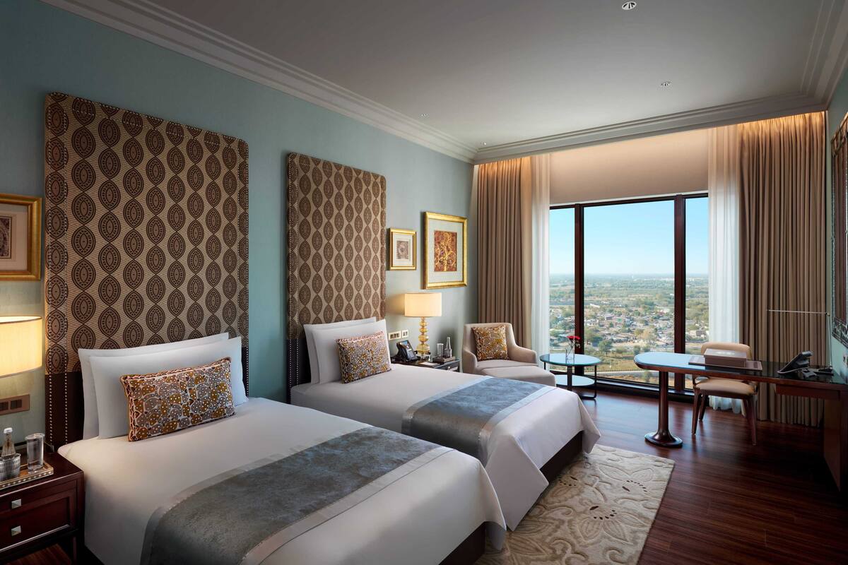 Ein Hotelzimmer mit zwei Betten, einem Schreibtisch, einem Stuhl, einem kleinen Tisch und einem großen Fenster mit Blick auf die Stadt.