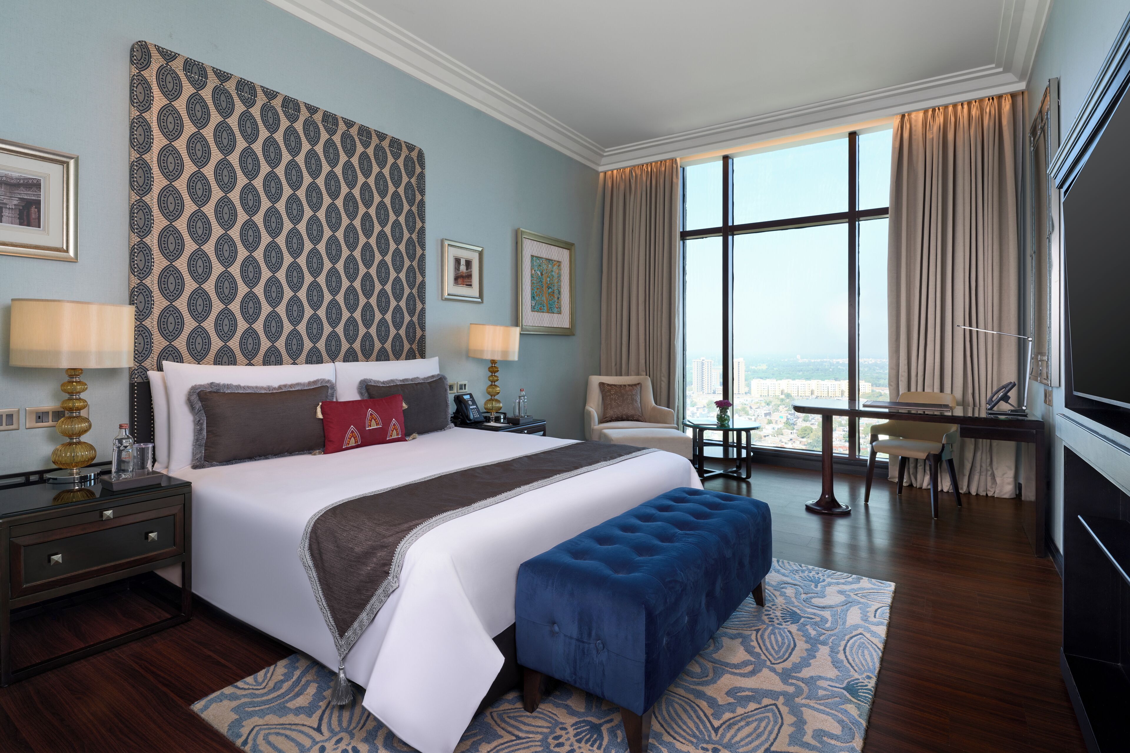 A hotel room with a large bed, a blue ottoman, a desk, and a view of the city.