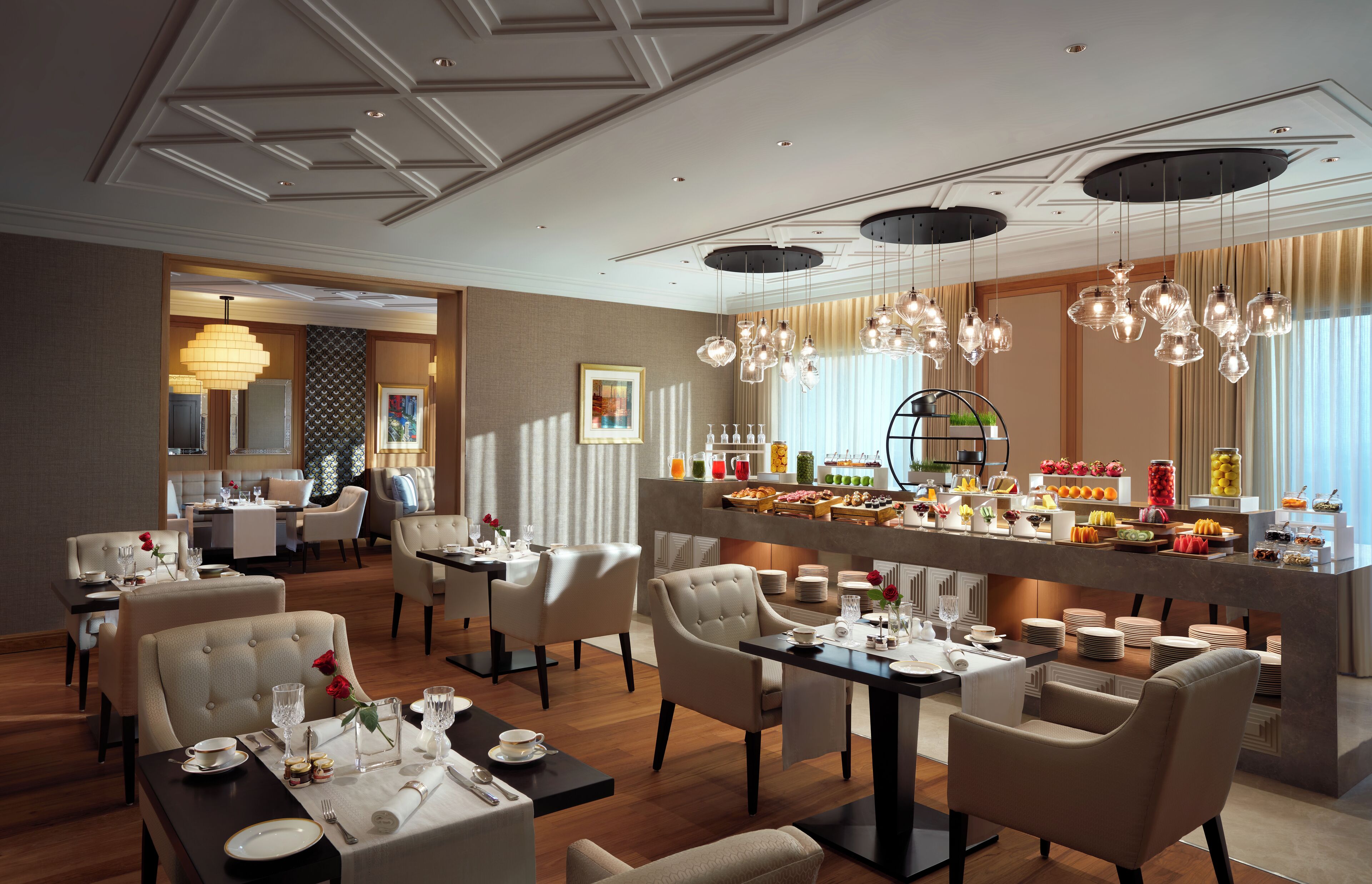 Un comedor de hotel con varias mesas y sillas, una barra de buffet y un techo bien iluminado con lámparas colgantes.