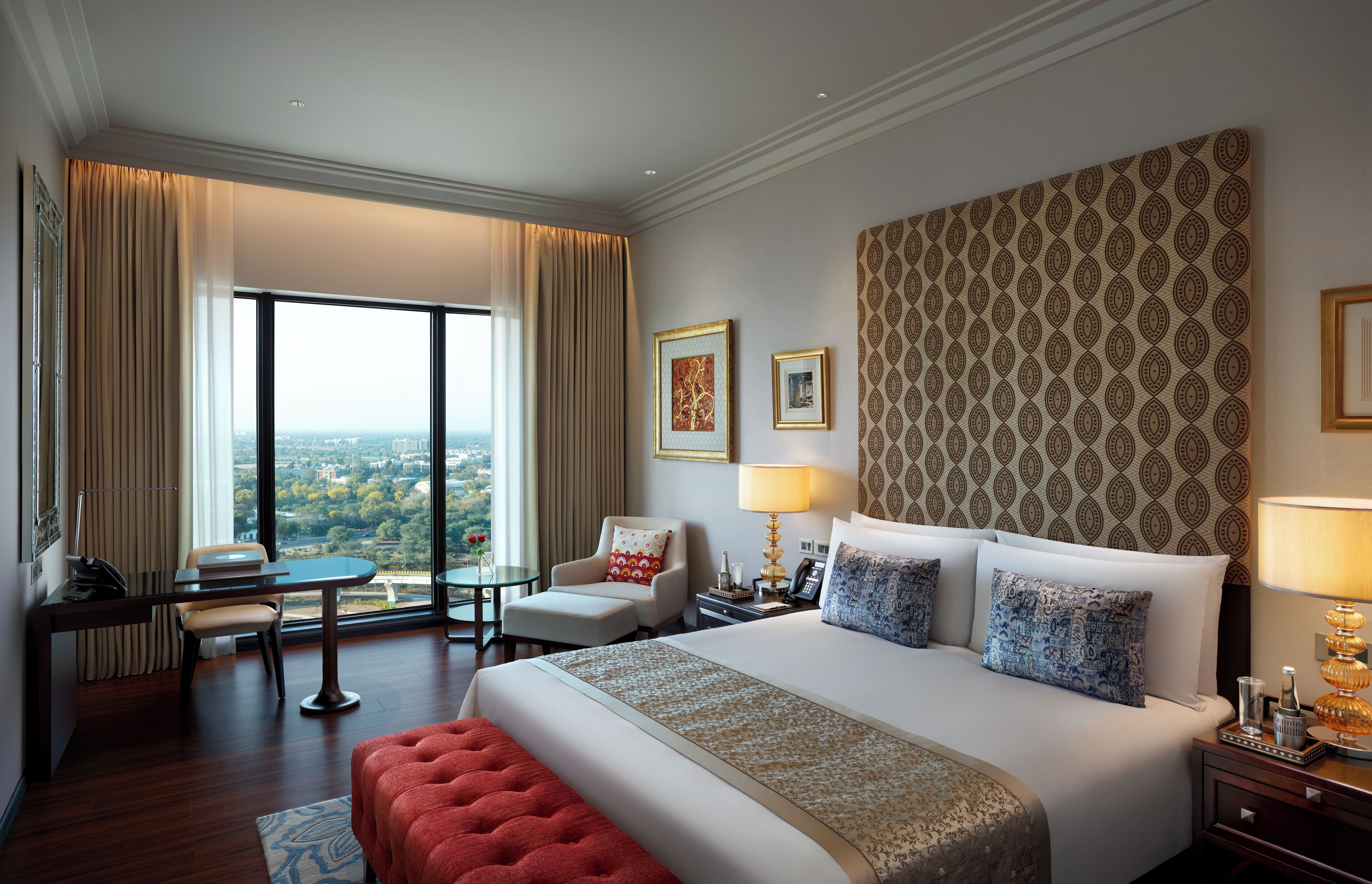 A hotel room with a large bed, a desk with a chair, a small table, and a view of the city through large windows.