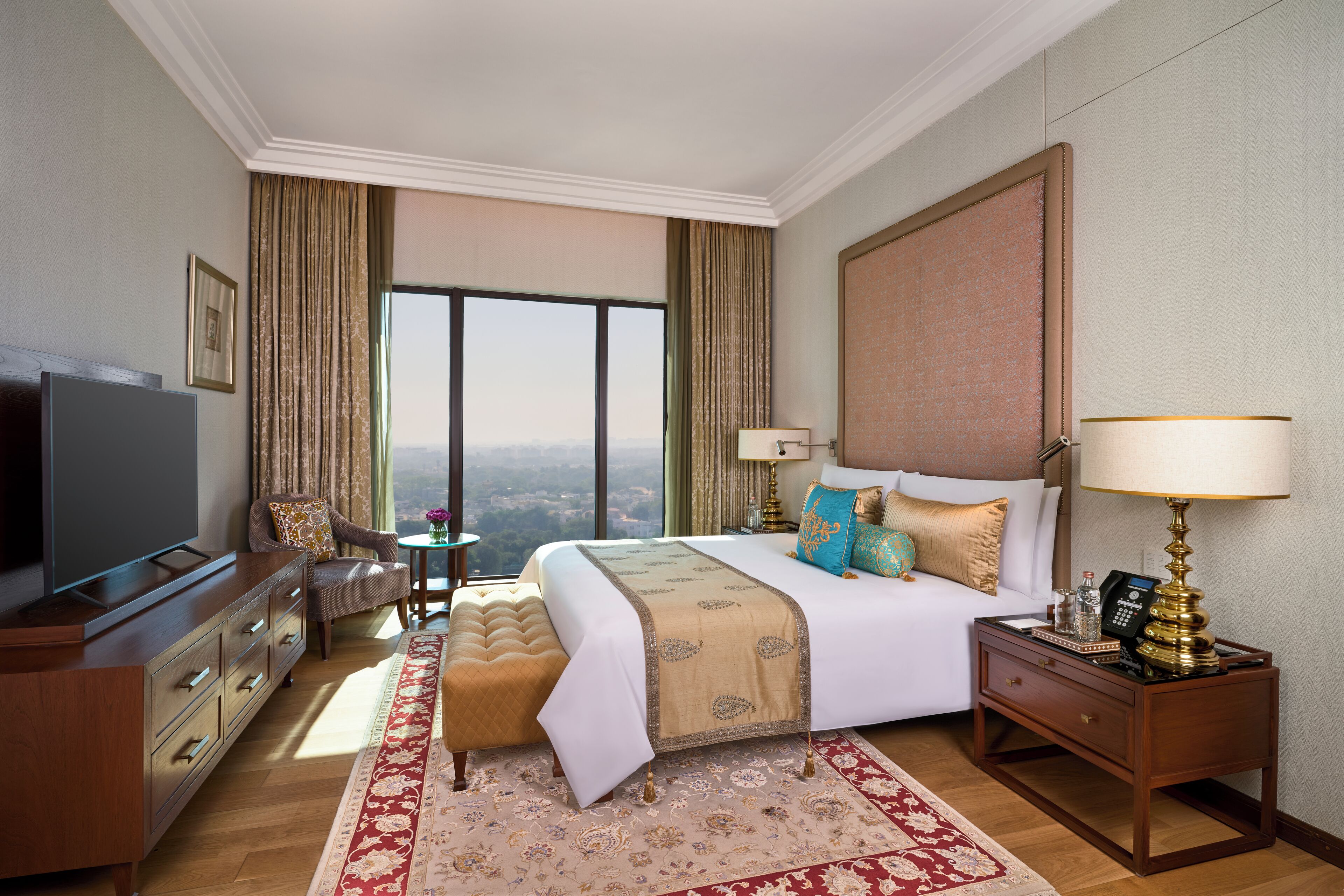 A hotel room with a large bed, a TV, a desk with a lamp, a chair, and a view of the city.