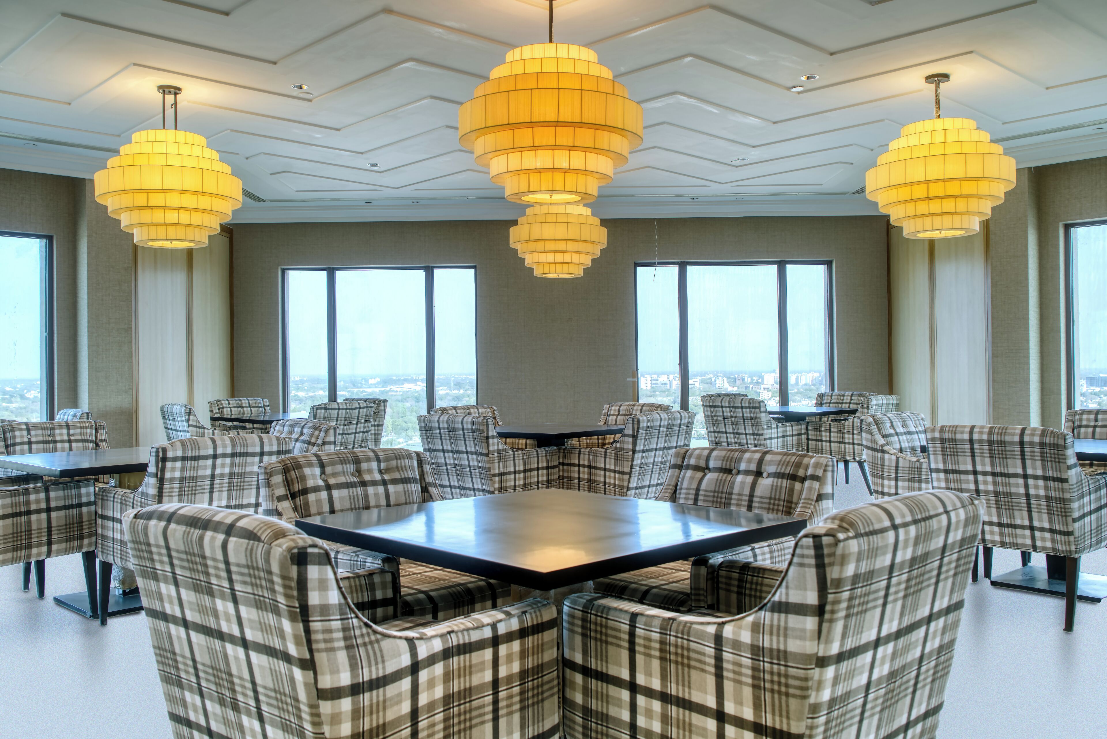 A conference room with a large table surrounded by plaid chairs, large windows, and three hanging chandeliers.