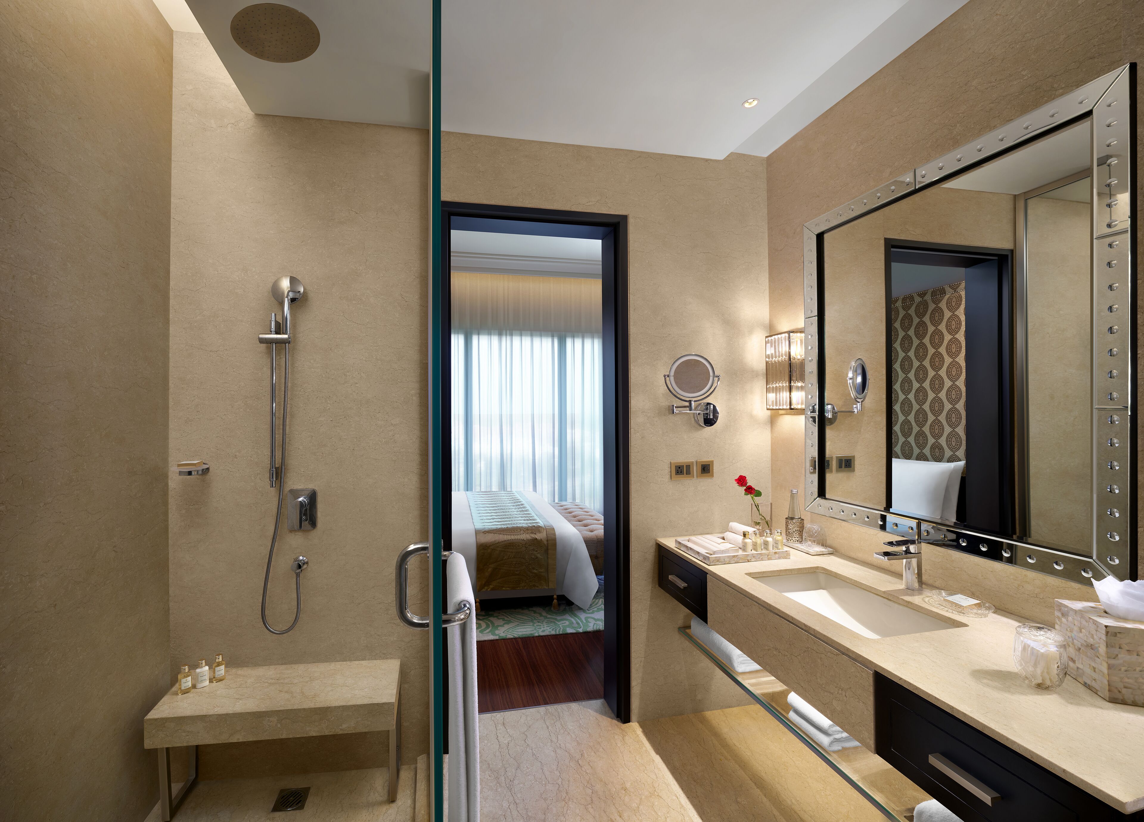 A modern bathroom with a glass shower enclosure, a vanity with a large mirror, and a bed visible through an open door.