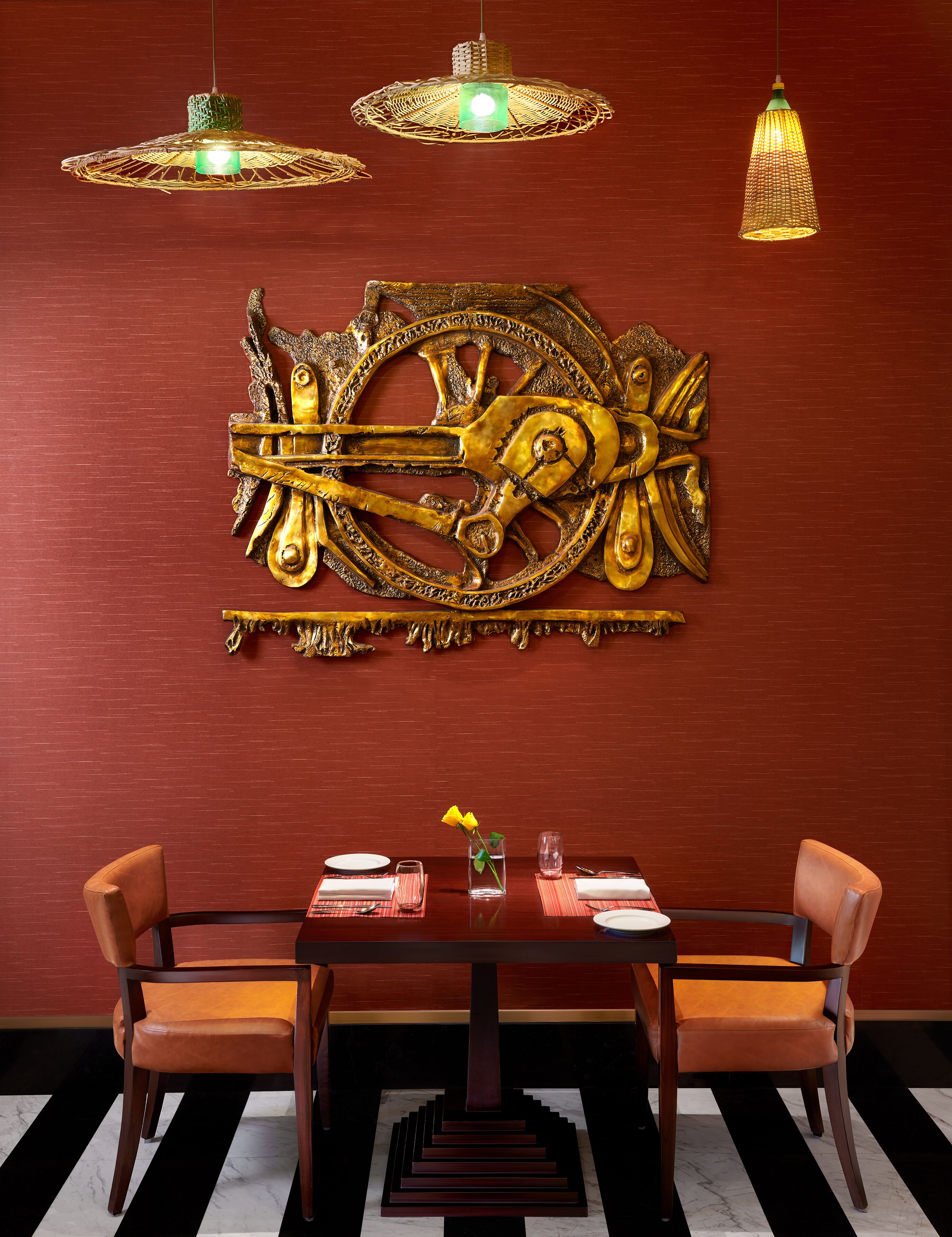 A dining area with a wooden table, two chairs, a decorative wall piece, and two hanging lights.