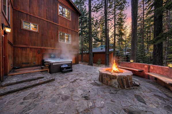 A wooden cabin with a stone patio, a hot tub, and a fire pit.