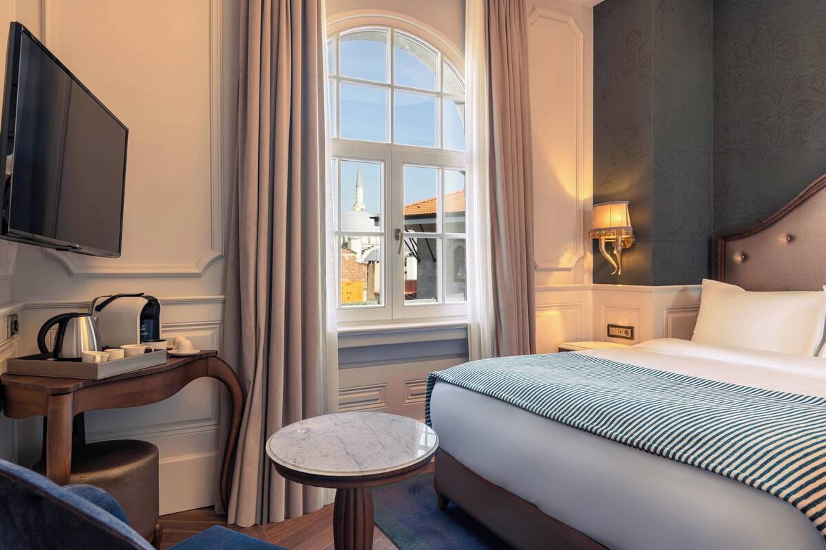 A hotel room with a large bed, a small round table, a desk with a flat-screen TV, a lamp, and a view of a building through a window.