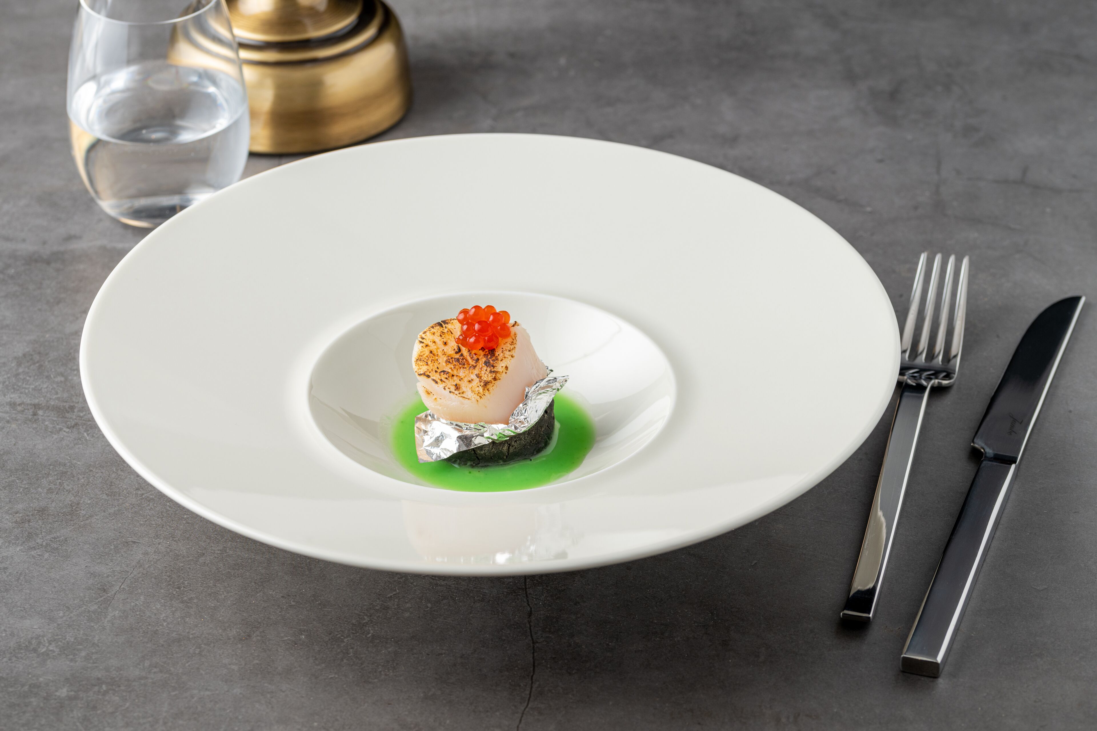Un plato blanco con un plato de mariscos, un vaso de agua y un objeto dorado al fondo.