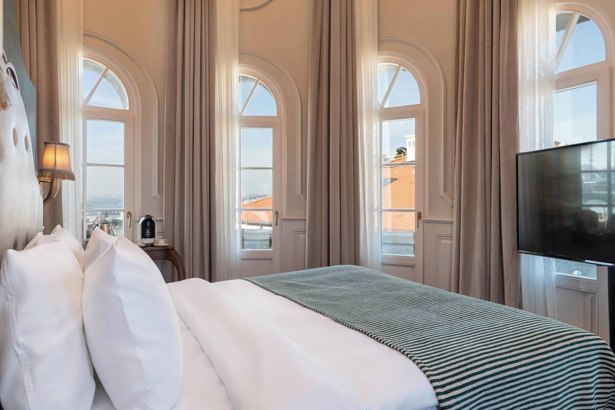 A hotel room with a large bed, a flat-screen TV, and a view of the city through arched windows.