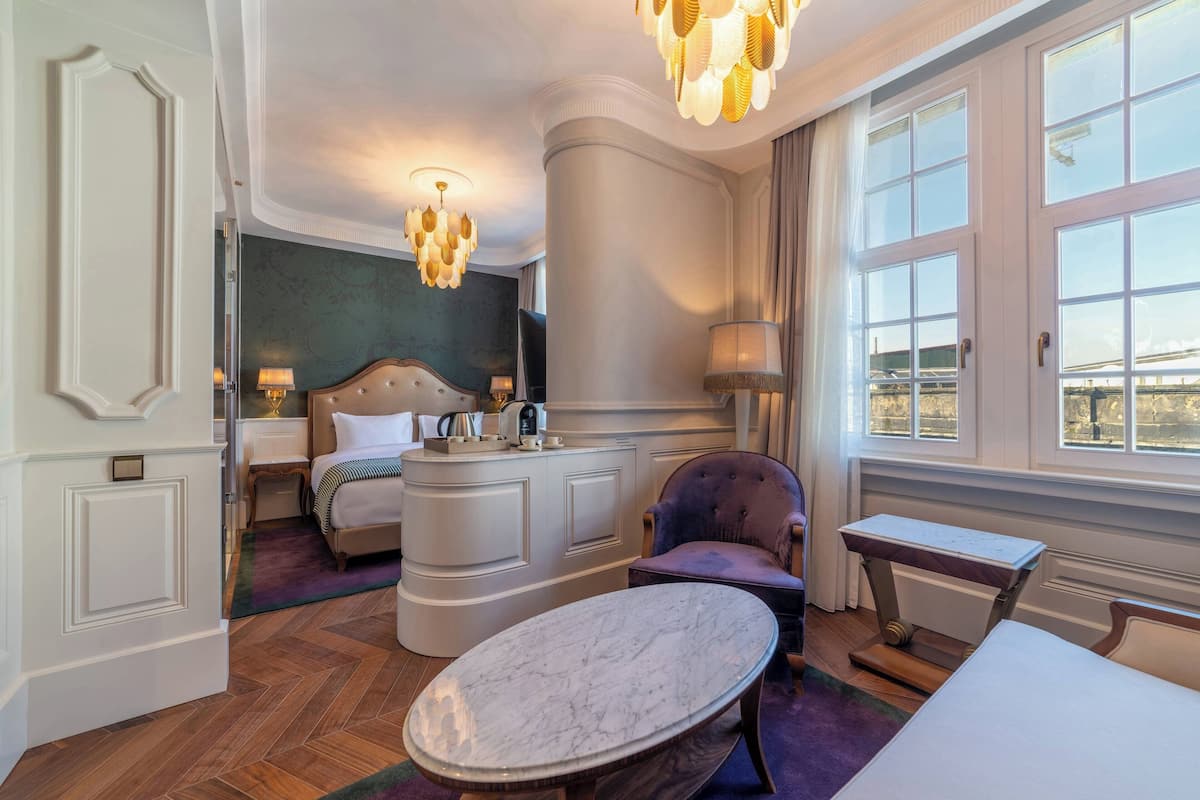 A hotel room with a bed, a vanity with a mirror, a chaise lounge, a marble table, and a large window with curtains.