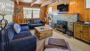 A cozy cabin interior with a wooden ceiling, a stone fireplace, a television mounted on the wall, and a leather sofa.