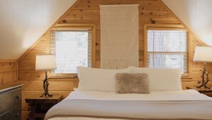 A cozy room with a wooden ceiling, a large bed with white bedding, two bedside tables with lamps, and a hanging macrame wall hanging.