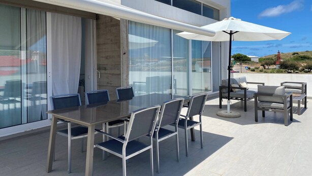 A modern outdoor dining area with a glass table and chairs, a white umbrella, and a view of the surrounding landscape. - Wonderful modern, spacious and comfortable villa (Fornells)
