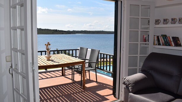 A room with a view of a lake, a wooden table with chairs, a sofa, and a bookshelf. - Apartment in Fornells (Fornells)