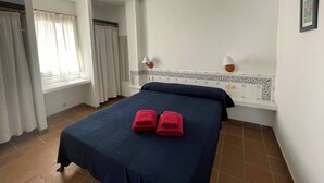 Une chambre d’hôtel avec un lit, deux lampes fixées au mur, une fenêtre avec des rideaux et un sol carrelé.