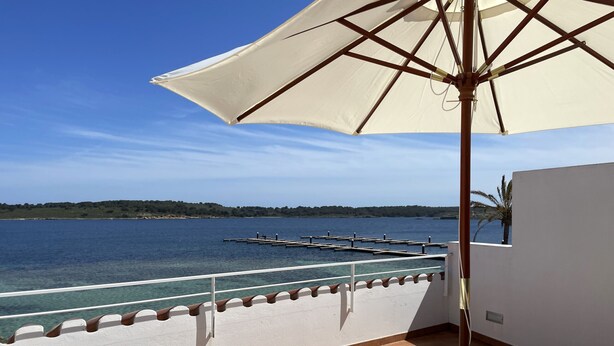 A balcony with two sunbeds, a large umbrella, and a view of the sea and a distant island. - Beautiful apartment in very front of the sea (Fornells)