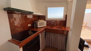A compact kitchen with a stove, microwave, and sink, a window with a view, and a balcony with a chair. - Beautiful apartment in very front of the sea (Fornells)