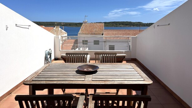 Een balkon met een houten tafel en stoelen, met uitzicht op een kustgebied met terracotta daken en een helderblauwe lucht.