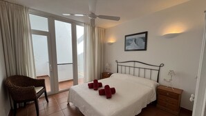 Een hotelkamer met een bed, een stoel, een nachtkastje, een ventilator en een raam met gordijnen.