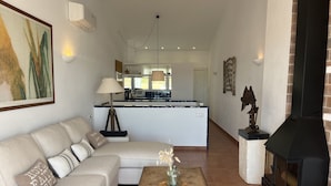 A living room with a sofa, a coffee table, a fireplace, and a kitchen in the background. - In a very quiet area within the town (Fornells)