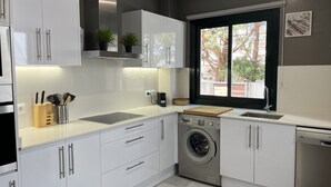 A modern kitchen with white cabinets, a built-in washing machine, and a window with a view of trees. - Superb luxury house in front of the sea (Fornells)