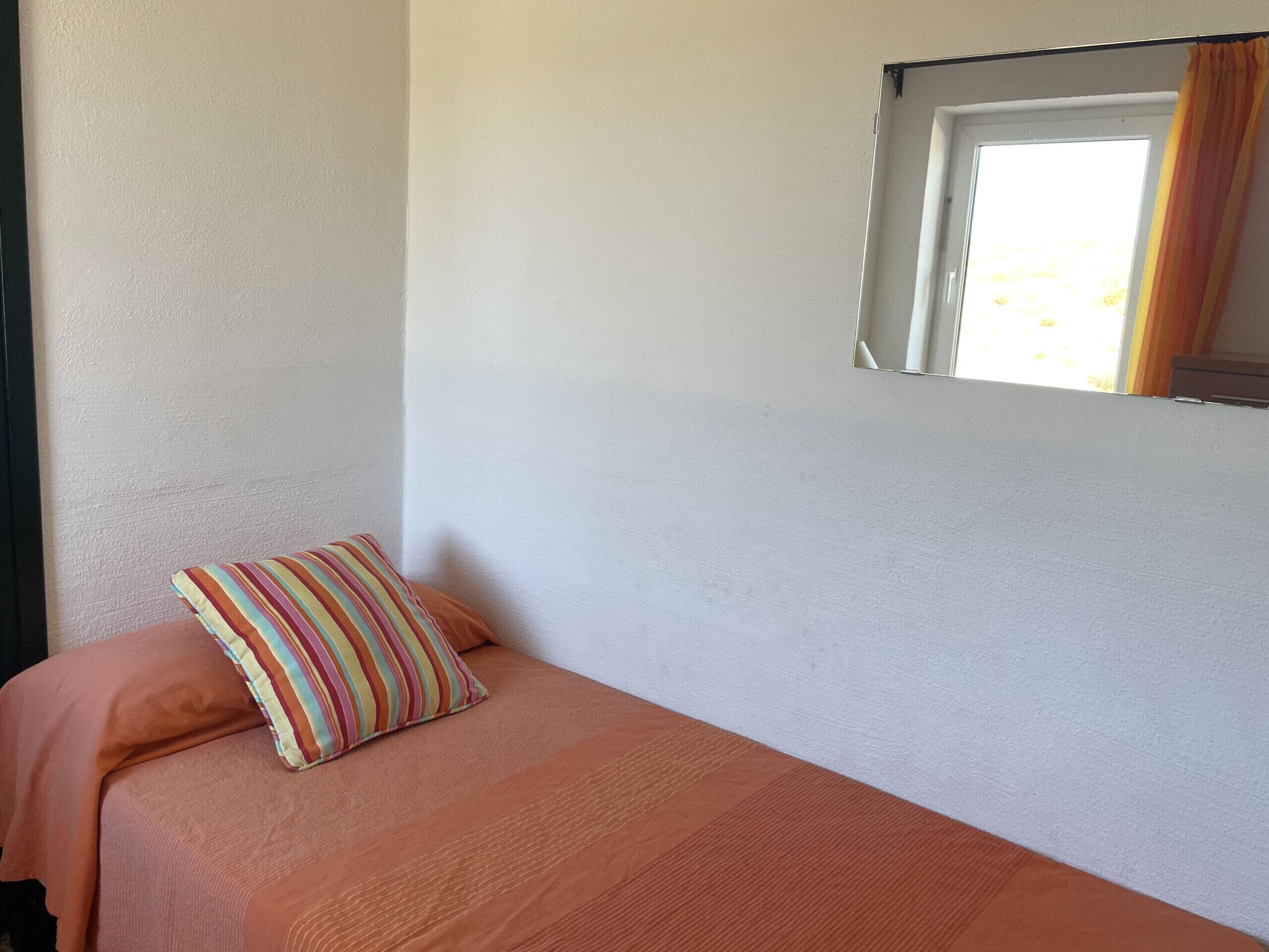 A single bed with a striped pillow and a plain orange bedspread in a room with white walls and a window with curtains.