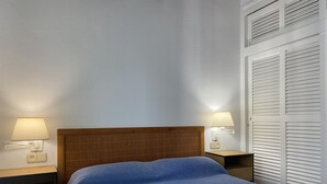 Habitación de hotel con cabecera de madera, colcha azul con textura, dos lámparas de noche y una ventana con contraventanas.