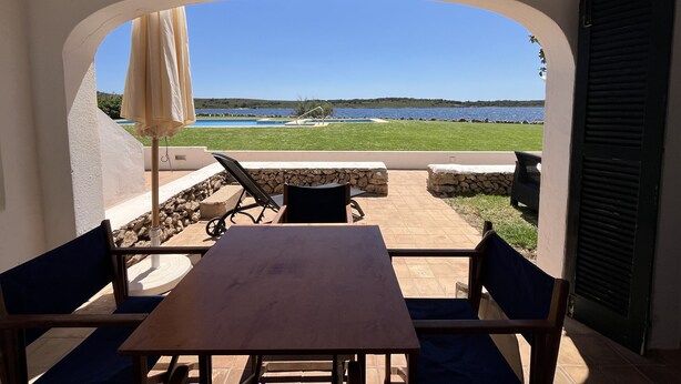 Un patio con una mesa y sillas, vista a una piscina y una playa, y un muro de piedra.