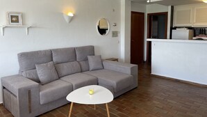 Un salon aux couleurs neutres, avec un canapé gris, une petite table blanche et une bougie allumée. On aperçoit une cuisine à l’arrière-plan, avec des armoires blanches et un plan de travail.