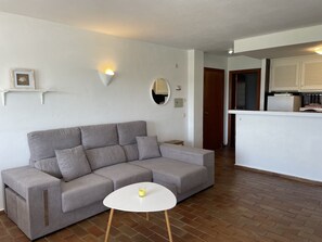 Un salon aux couleurs neutres, avec un canapé gris, une petite table blanche et une bougie allumée. On aperçoit une cuisine à l’arrière-plan, avec des armoires blanches et un plan de travail.
