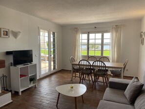 Un salon comprenant un téléviseur, une table à manger avec des chaises, un canapé et une vue sur une étendue d’eau grâce à de grandes fenêtres.