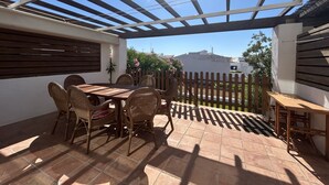 An outdoor patio with a pergola, tiled flooring, wicker furniture, and a view of a white building in the background.