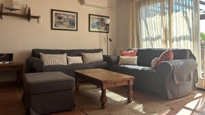 A living room with a sofa, ottoman, and wooden coffee table. There are two framed pictures on the wall, a lamp, and a window with curtains.