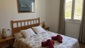 Een slaapkamer met een houten hoofdbord, een bloemenbedekking, twee witte kussens, een houten nachtkastje met een lamp, een lijst met een afbeelding van zeilboten, en een raam met gordijnen.