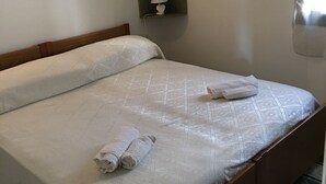 Un lit bien fait, avec une couette blanche et deux serviettes enroulées, une lampe de chevet et une petite étagère ornée d’un objet décoratif.