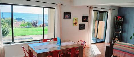 Un comedor con mesa y sillas, ventanas amplias con vista al mar y una estantería.