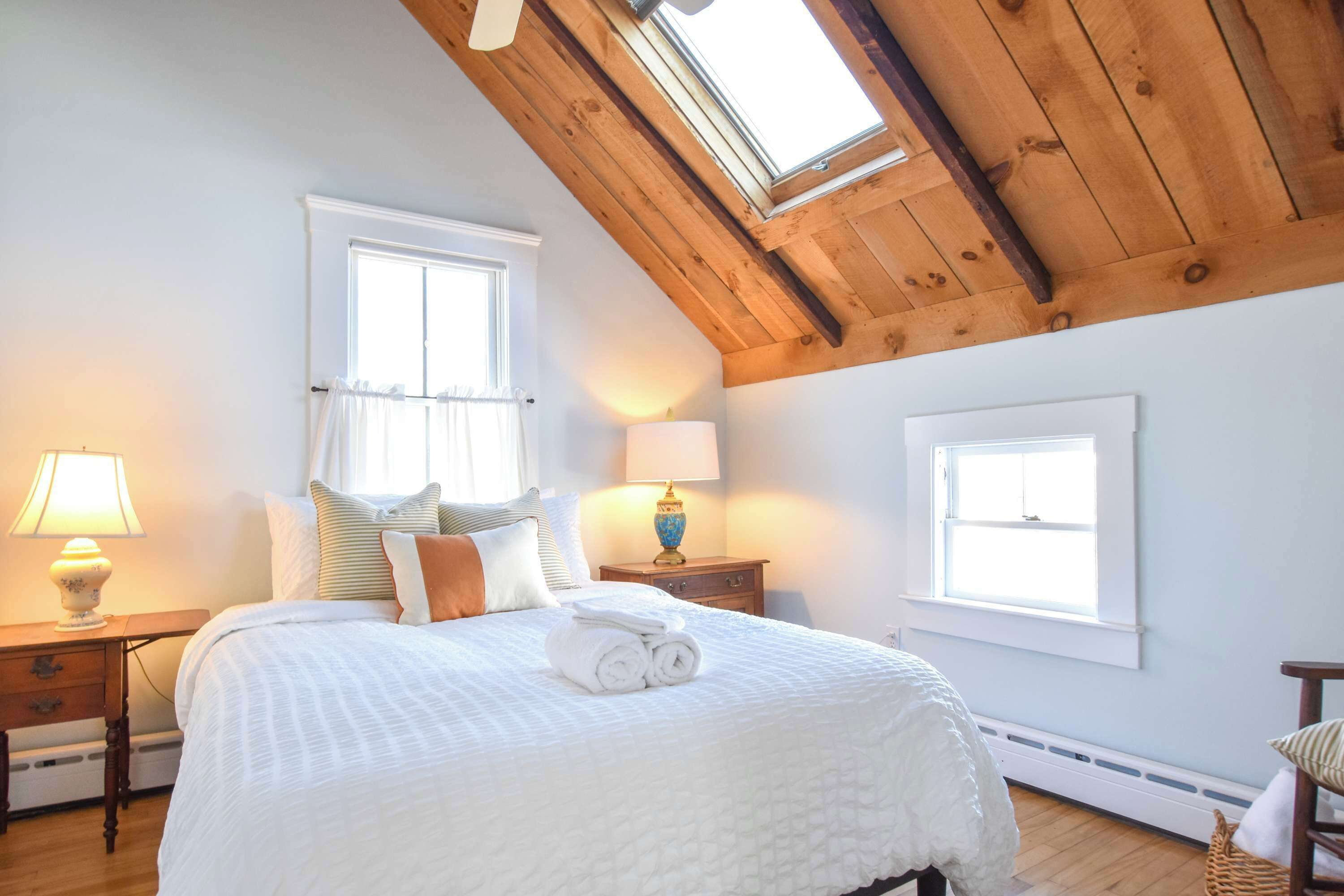 Ein Schlafzimmer mit schrägverdeckter Decke, zwei Fenstern, einem Bett mit einem zusammengerollten Handtuch, einem Nachttisch mit Lampe und einem Holzboden.