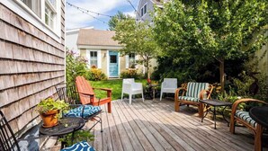 A wooden deck with outdoor furniture, including chairs and a table, surrounded by lush greenery and a house in the background. - 5 Min Walk to Town, Patio, Yard, AC! (Provincetown)