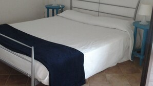 Un lit simple avec un matelas blanc et une couverture bleu foncé, flanqué de deux tables de chevet bleues avec des lampes, dans une pièce dont le sol est recouvert de carreaux.