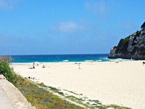 A beach with clear blue water, a rocky cliff, and a few people.