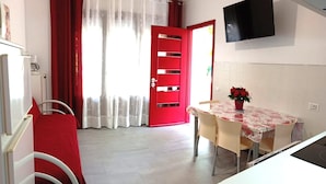 A room with a red door, a dining table with chairs, a potted plant, and a flat-screen TV mounted on the wall. - splendid two-room apartment with pergola (Bibione)
